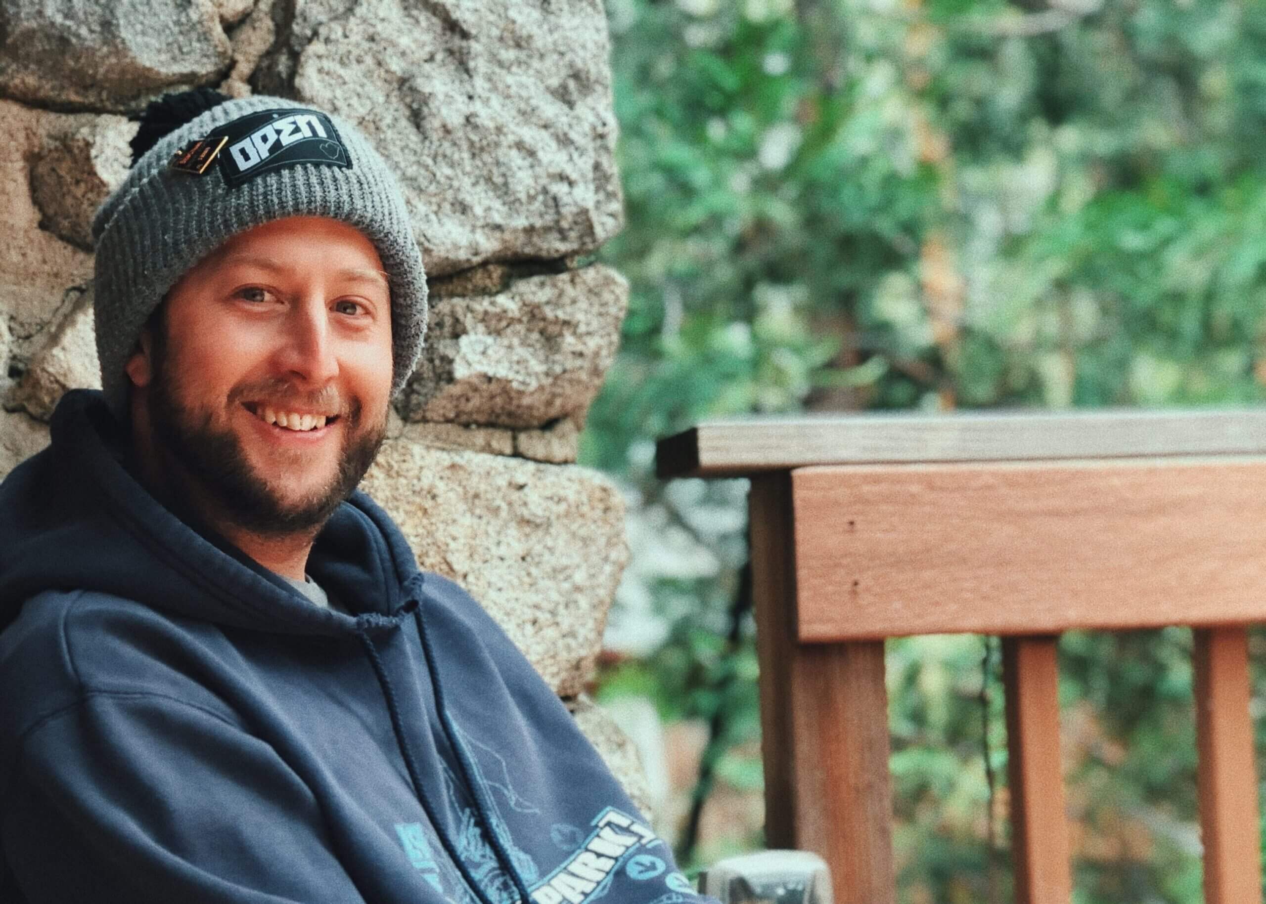 Smiling man wearing a gray knit beanie and navy hoodie, leaning against a stone wall beside a wooden railing outdoors.