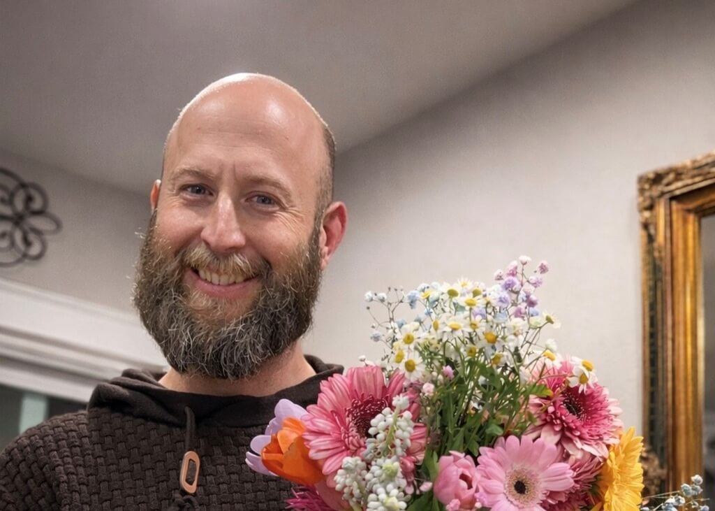 Smiling man with a beard holding a colorful bouquet of pink, yellow, and white flowers indoors.