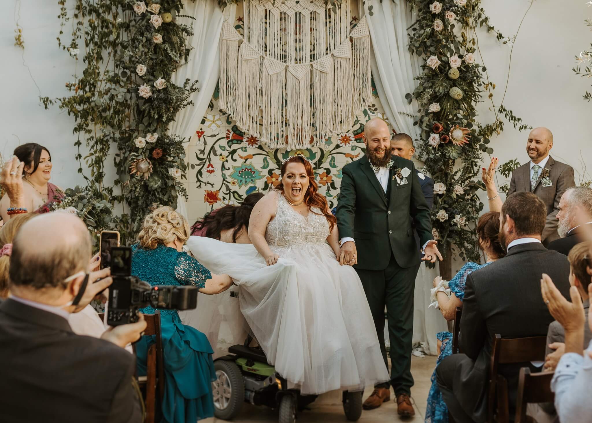 Bride and groom walk down the aisle, smiling as guests cheer in a bohemian, floral wedding backdrop with macrame fringe.
