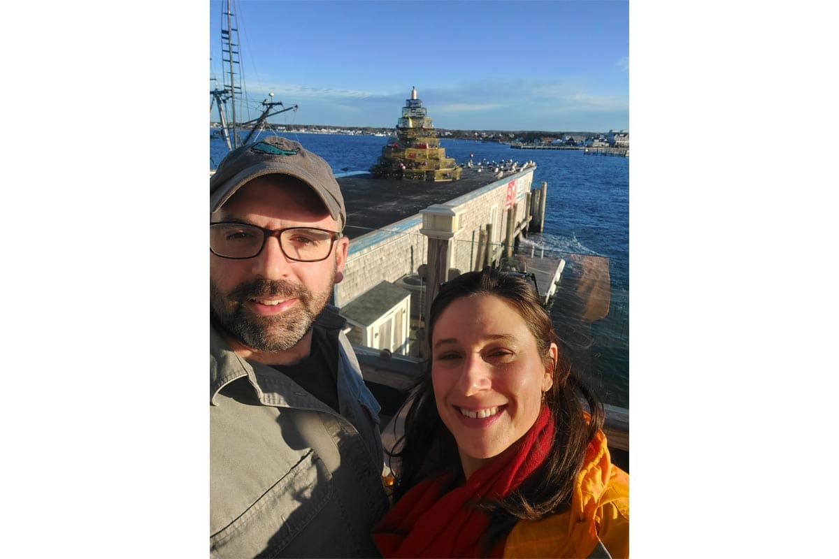Two people smiling for a selfie on a harbor pier, with the ocean and a tiered sculpture in the background.