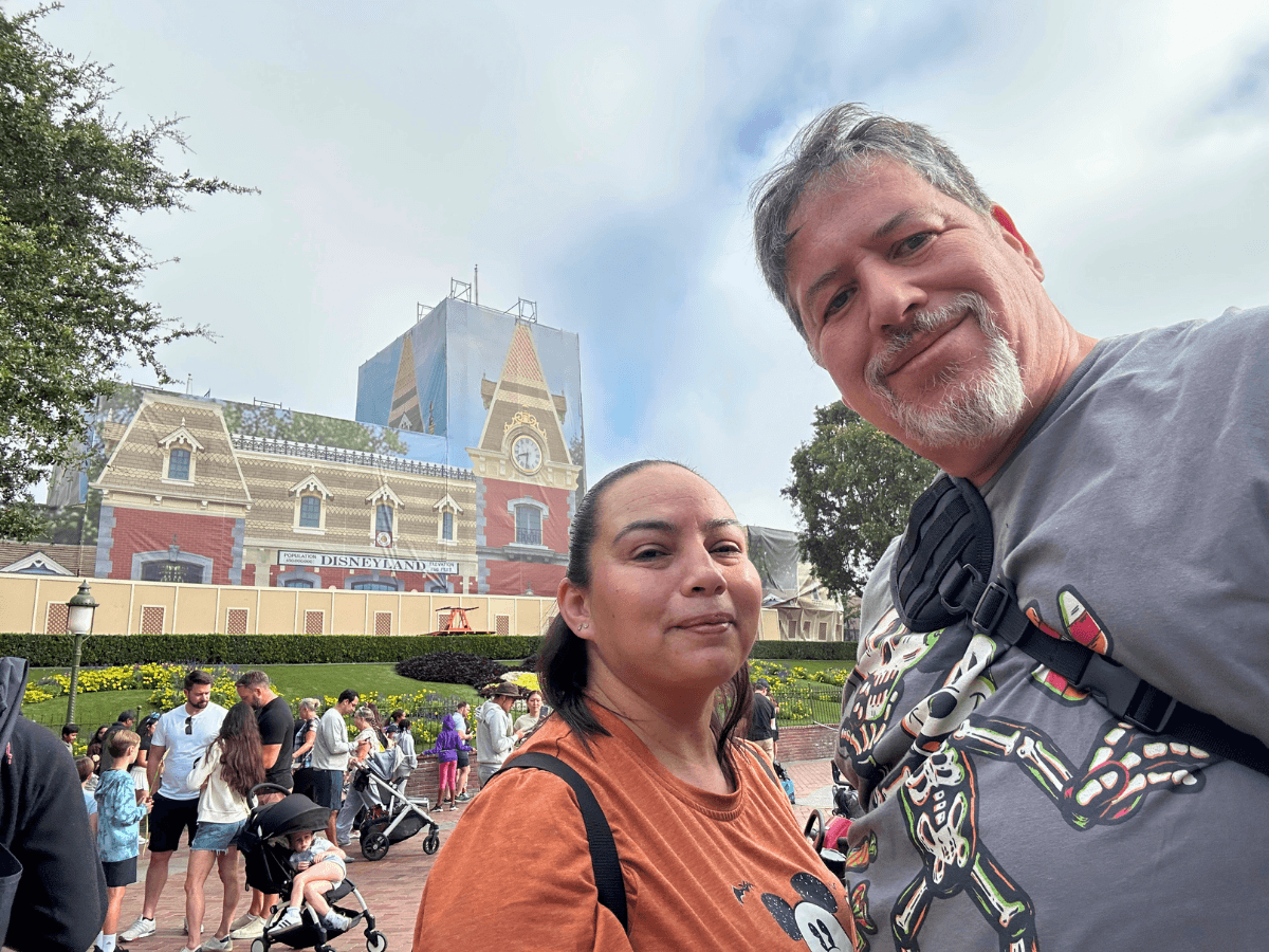 A couple poses for a selfie in front of the Disneyland entrance with a crowd and strollers behind them.
