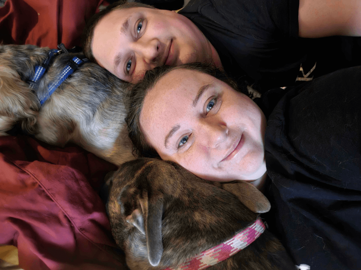 Two people cuddling on a red couch, smiling, with a brown dog wearing a pink plaid collar resting between them on the couch