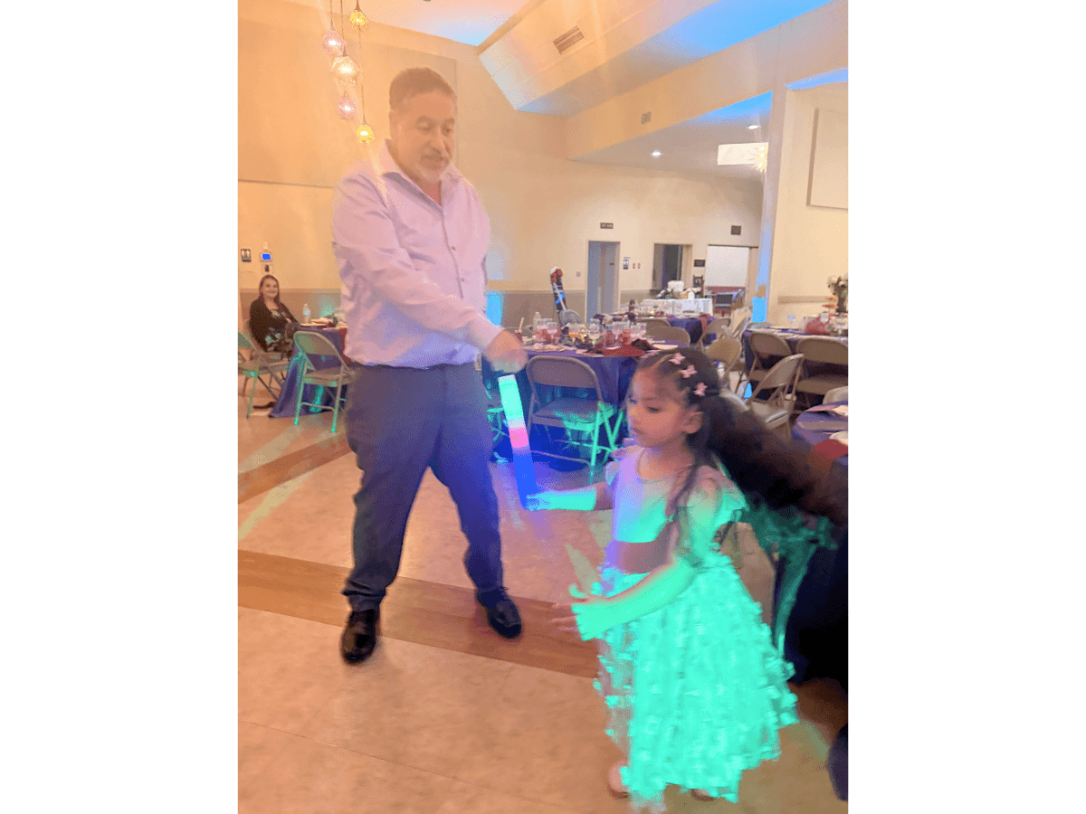 A man in a dress shirt dancing with a young girl holding a glowing blue-green baton at a party venue.