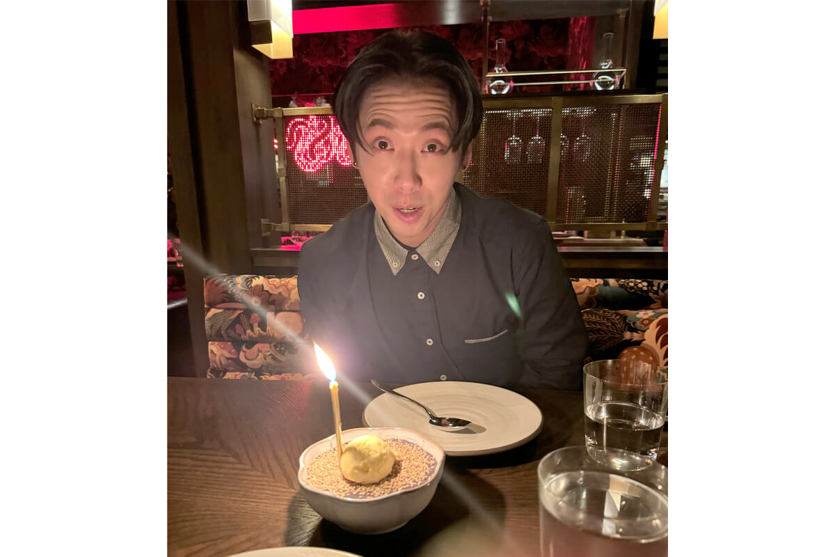 Man at a restaurant table with a lit birthday candle on a dessert bowl in front of him, smiling at the camera.