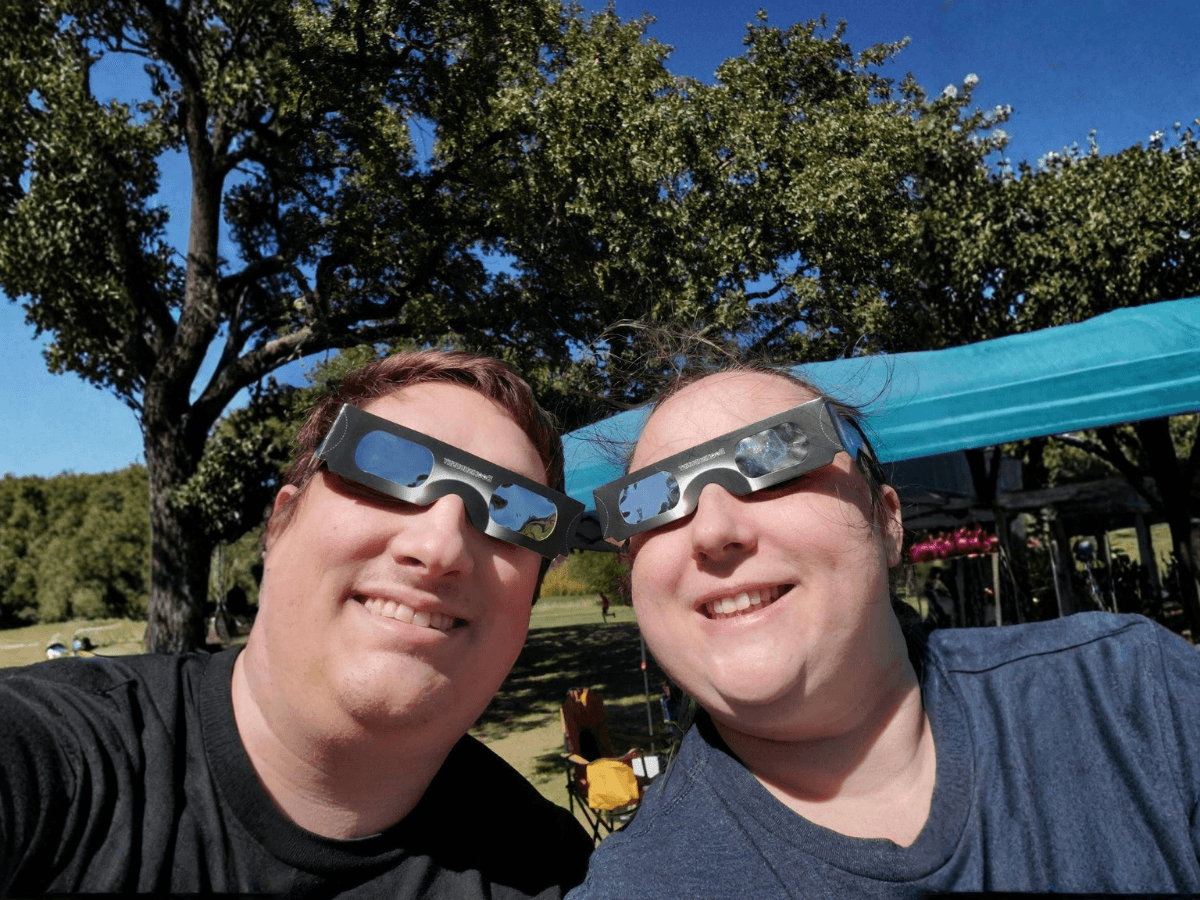 Two friends wearing solar eclipse glasses take a sunny park selfie, smiling at the camera.