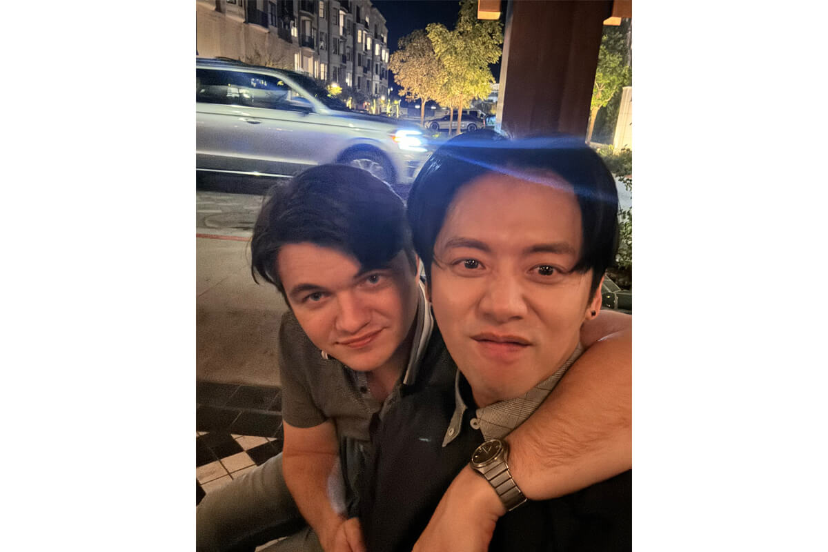 Two friends pose for a nighttime selfie on a city sidewalk, one with his arm around the other, smiling at the camera in a casual street scene.