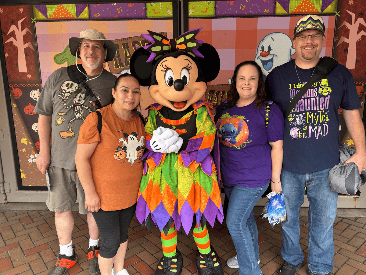 Four adults posing with Minnie Mouse in a bright harlequin costume at a Halloween-themed backdrop, all smiling at the camera.