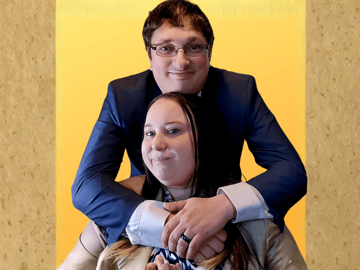 Couple posing together: a man in a dark suit hugs a smiling woman from behind, indoors against a yellow backdrop.