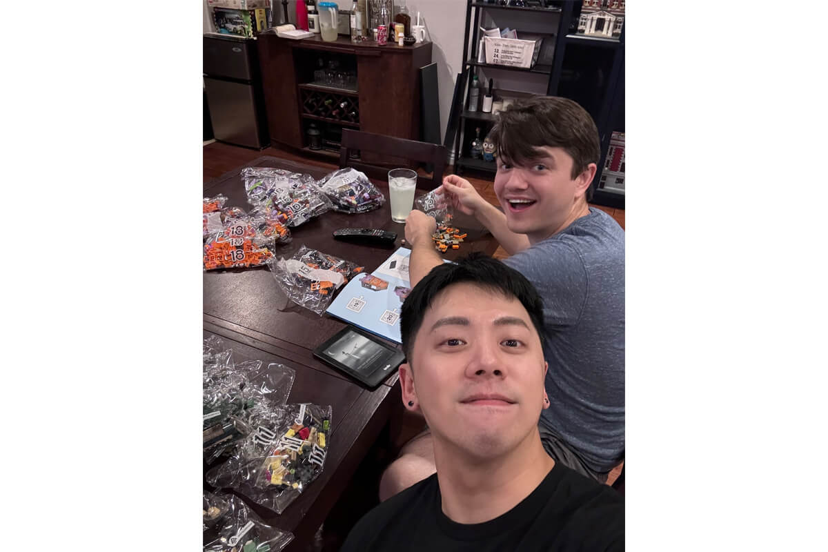 Two men sit at a table with open candy bags and game pieces, one taking a selfie while the other smiles at the camera in a home setting.