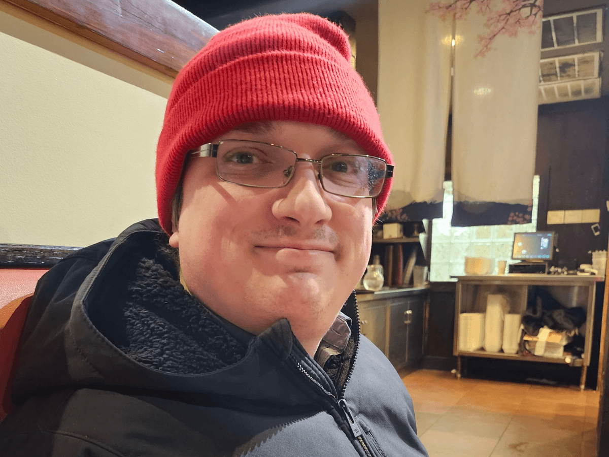 Person wearing a red knit beanie and glasses, smiling at the camera in a cafe or restaurant setting.