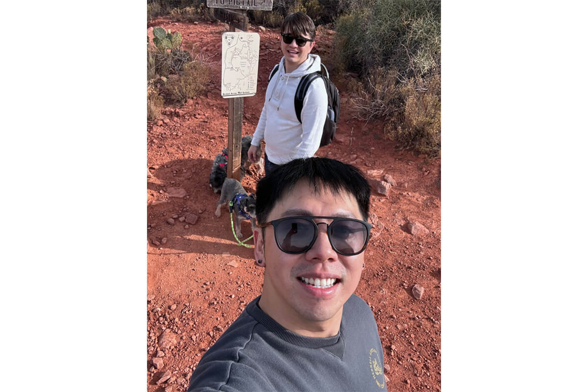 Selfie of two hikers on a red dirt trail; man in the foreground wears sunglasses and a gray shirt, with two dogs on leashes behind him.