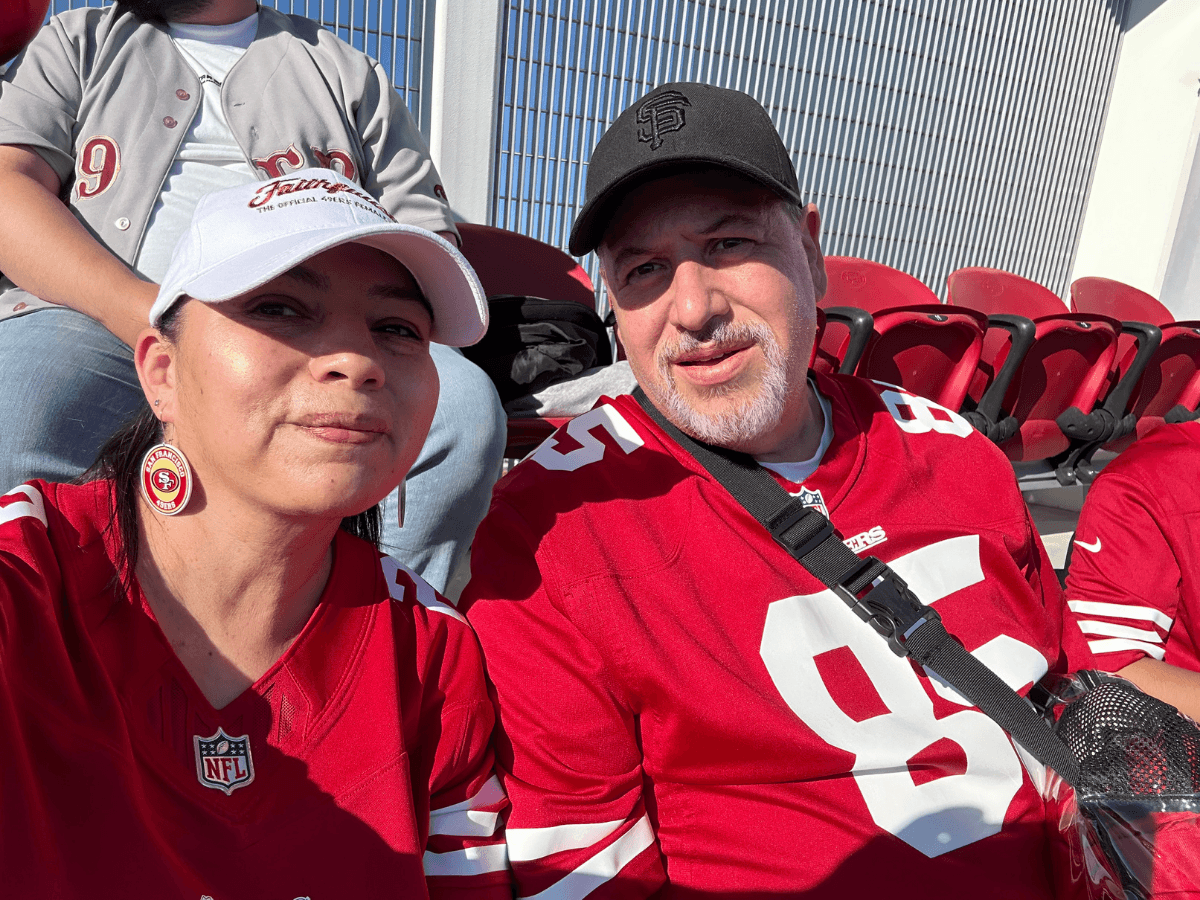 Two football fans in San Francisco 49ers jerseys posing for a selfie in a stadium with red seats behind them.