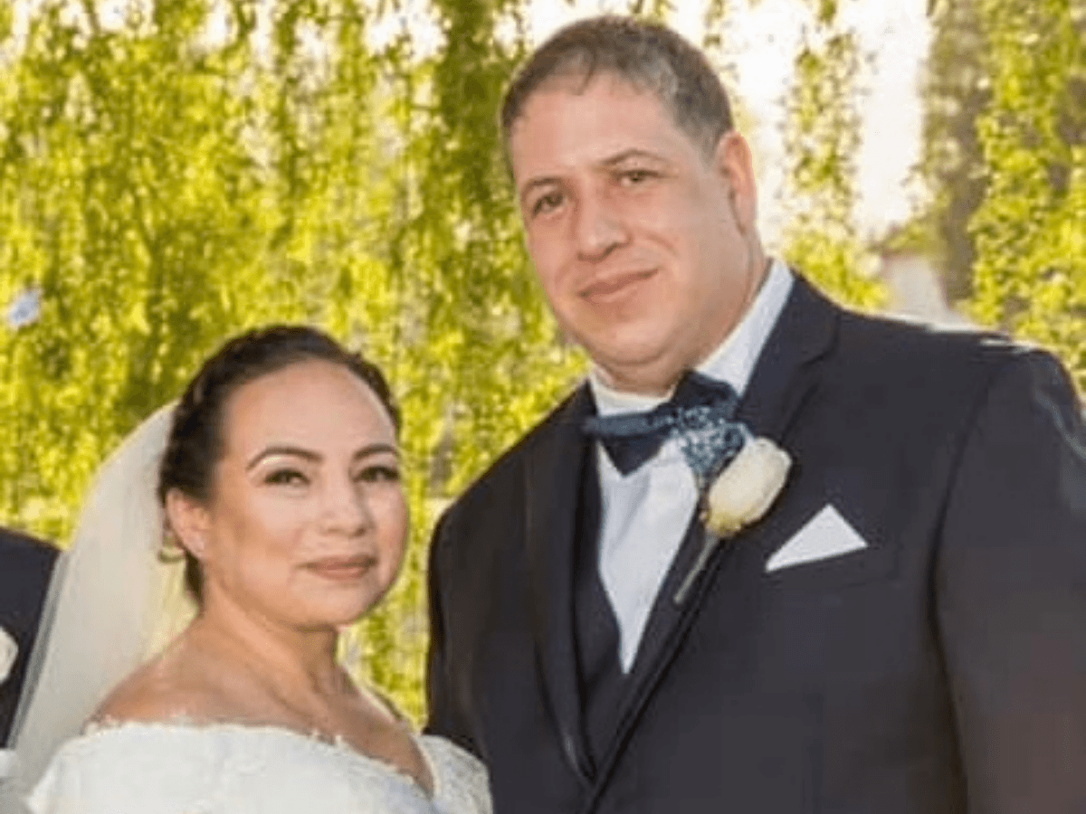 Bride in a white wedding gown and a groom in a black tuxedo pose together outdoors with greenery in the background.