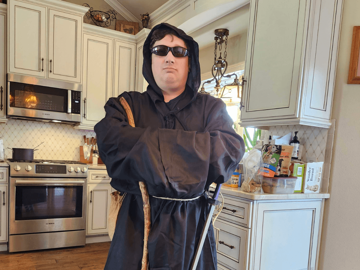 Person in a black hooded robe and sunglasses stands in a bright kitchen, gripping a wooden staff with a sword at the belt."