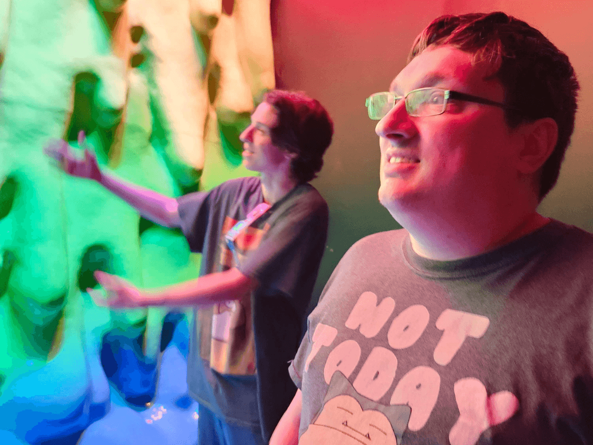 Two people explore a bright, colorful wall exhibit with neon green and pink lighting; one man in foreground smiles at the wall.