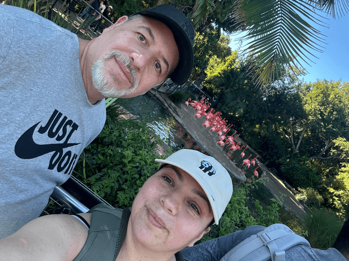 Two adults taking a selfie outdoors with a line of pink flamingos by a pond in the background.