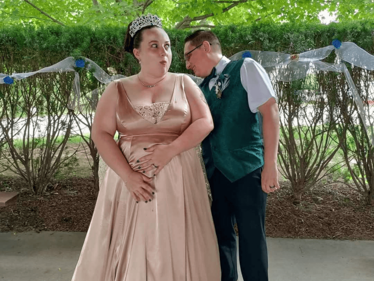 Bride in a peach gown with a tiara posed with a man in a green vest outdoors, with decorative ribbon in the background.
