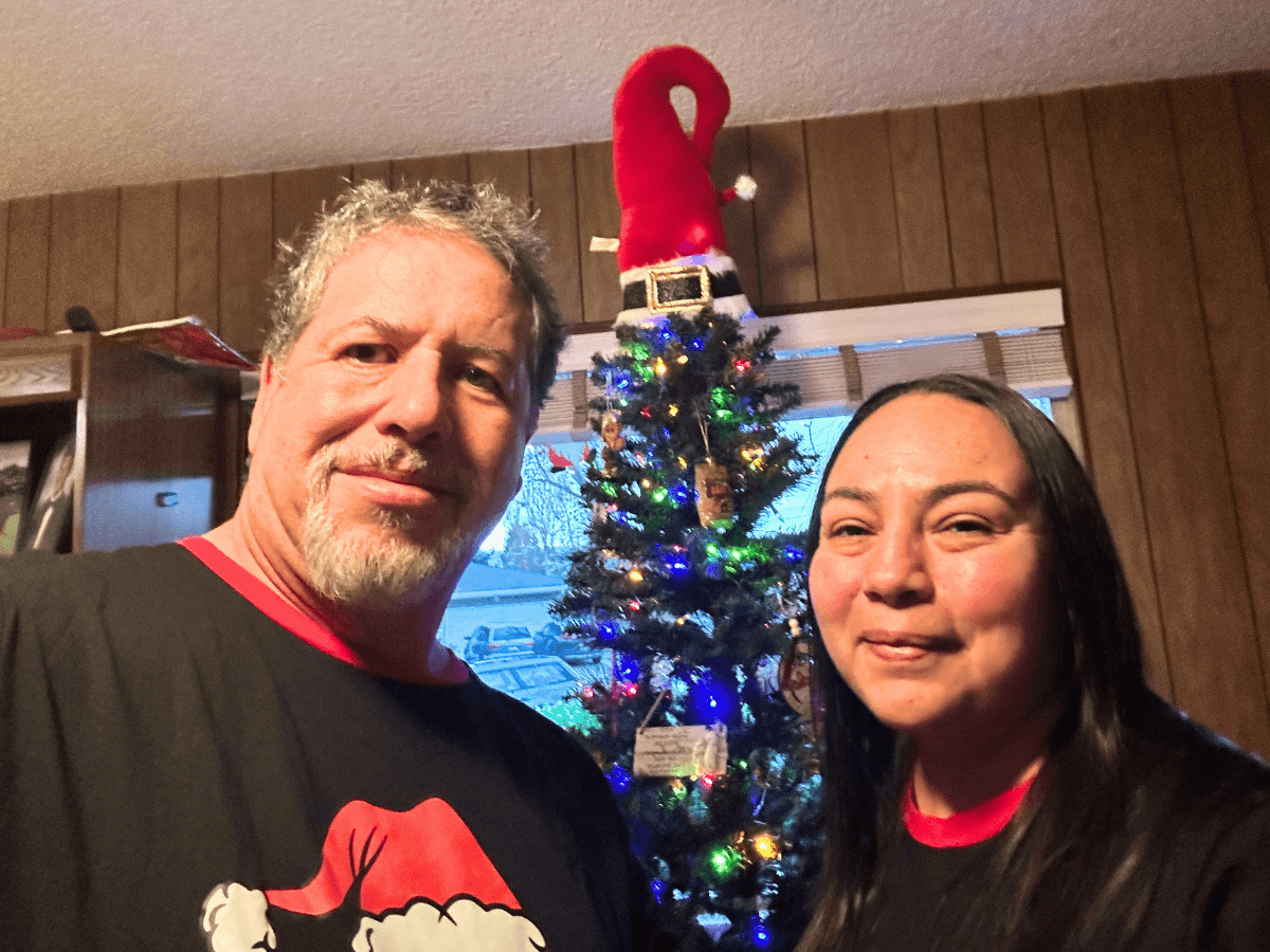 Man and woman pose in a living room with a Christmas tree topped by a Santa hat behind them, smiling at the camera.