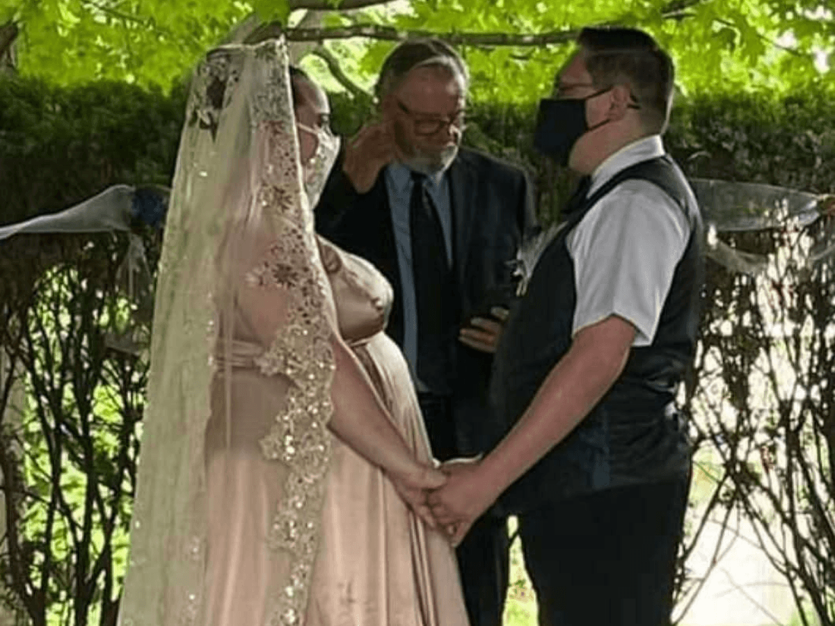 Pregnant bride in a lace gown and veil holds hands with groom in a vest during an outdoor wedding ceremony with an officiant nearby.
