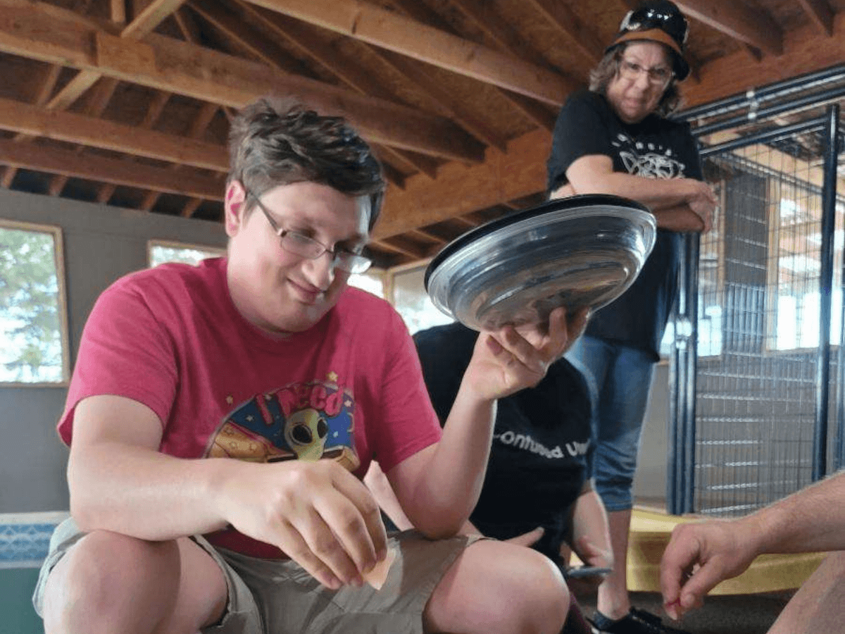 Young man in a red shirt balances a shiny metal bowl on his hand during a group activity indoors.