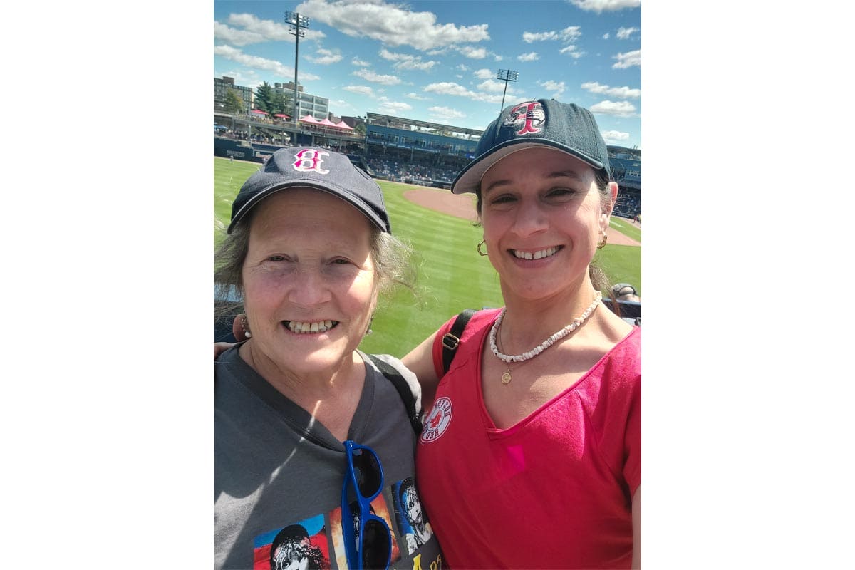 Two women at a baseball stadium on a sunny day, smiling for a selfie with the field in the background.
