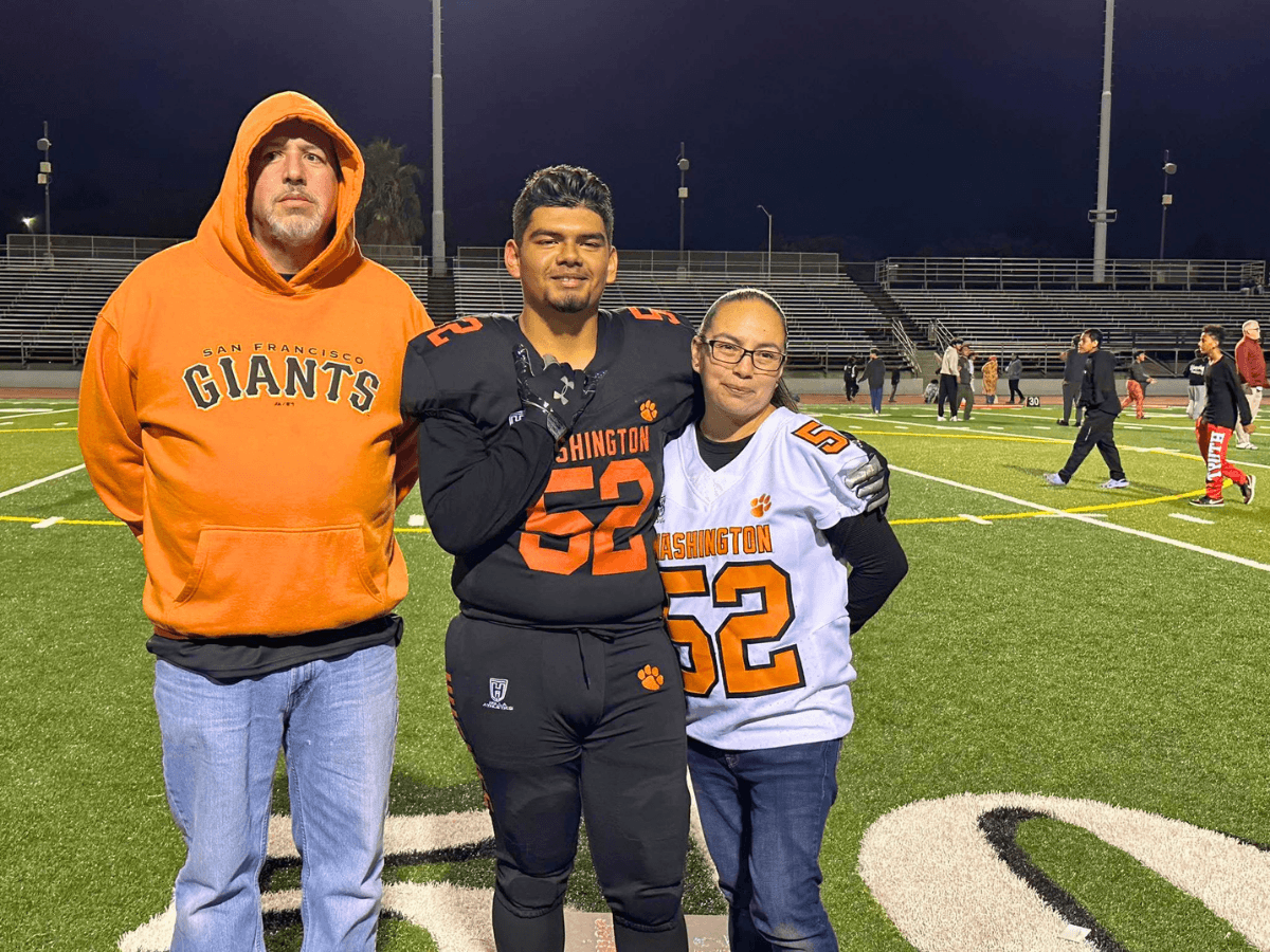 Three people posing together on a football field at night: man in orange hoodie, and two players in team jerseys (52) with arms around each other.