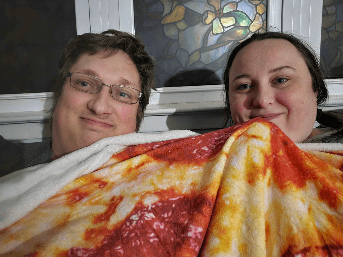 Two people snuggling under a colorful pizza-pattern blanket and smiling at the camera, with a stained-glass window behind them in a home setting.