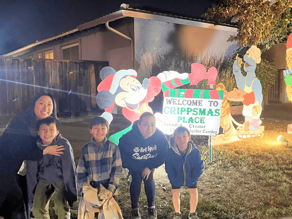 Family group posing at night in front of a Christmas display with a 'Welcome to Crippsmas Place' sign and cartoon cutouts, smiling for the camera.