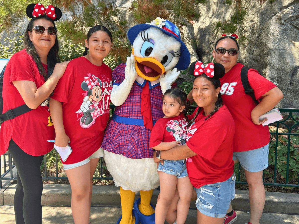 A group of five women and a young girl pose with a costumed Daisy Duck character outdoors, all wearing red shirts with Minnie Mouse themes.