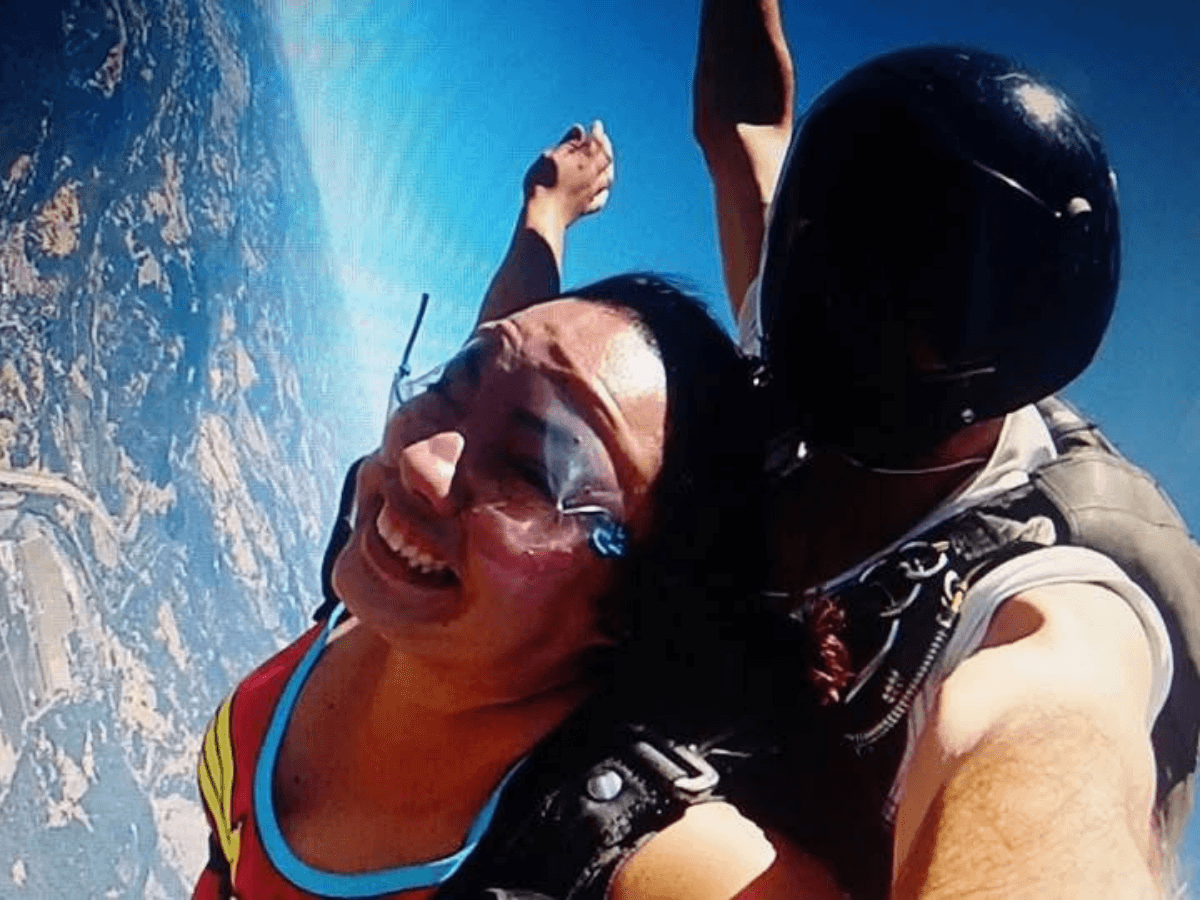 Tandem skydiving pair: a smiling woman in a red suit and goggles attached to a helmeted instructor against a clear blue sky and landscape below.
