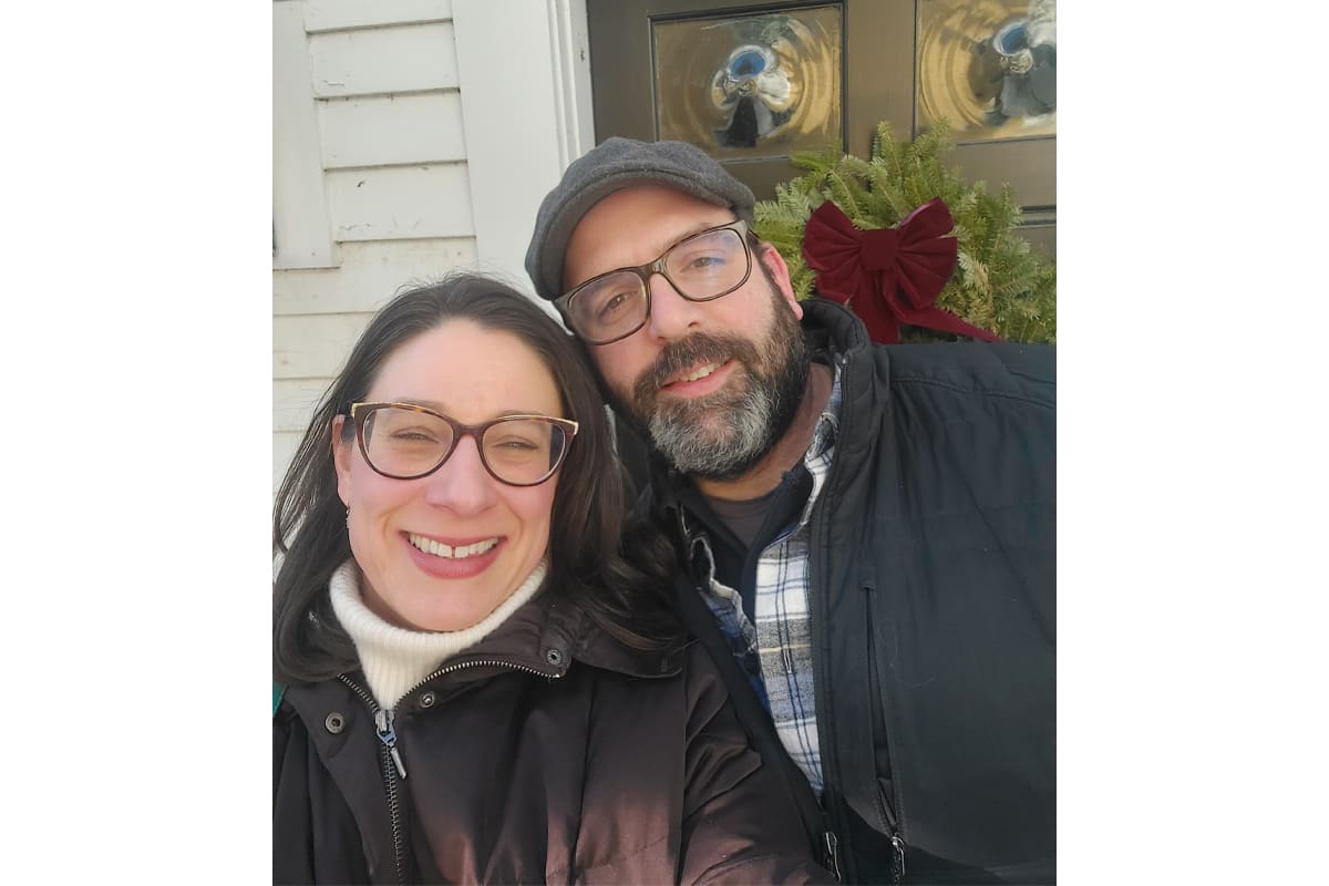 Smiling couple wearing glasses pose for a selfie outside a house with a wreath on the door? on winter coats and hats (informative description).