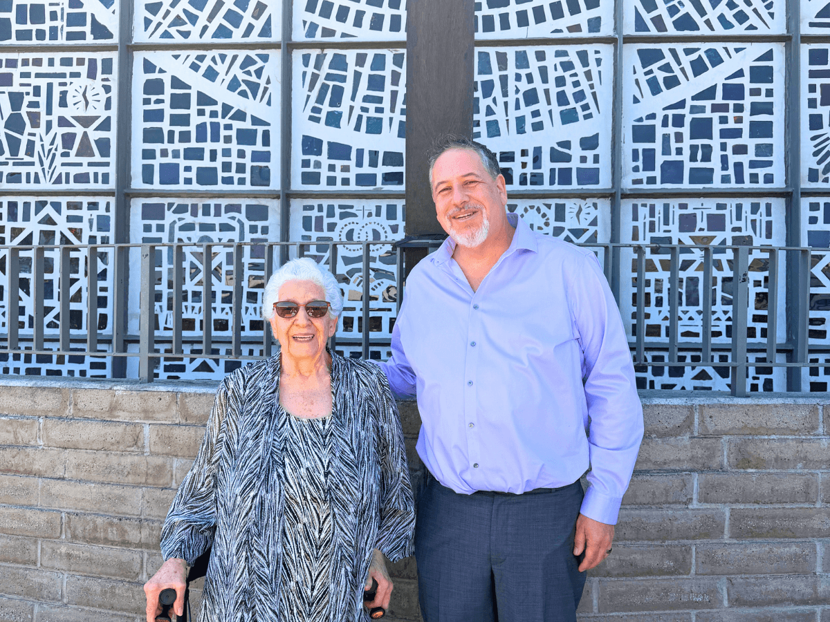 Smiling elderly woman with a cane and sunglasses poses with a man in a lavender shirt in front of a blue decorative patterned wall.