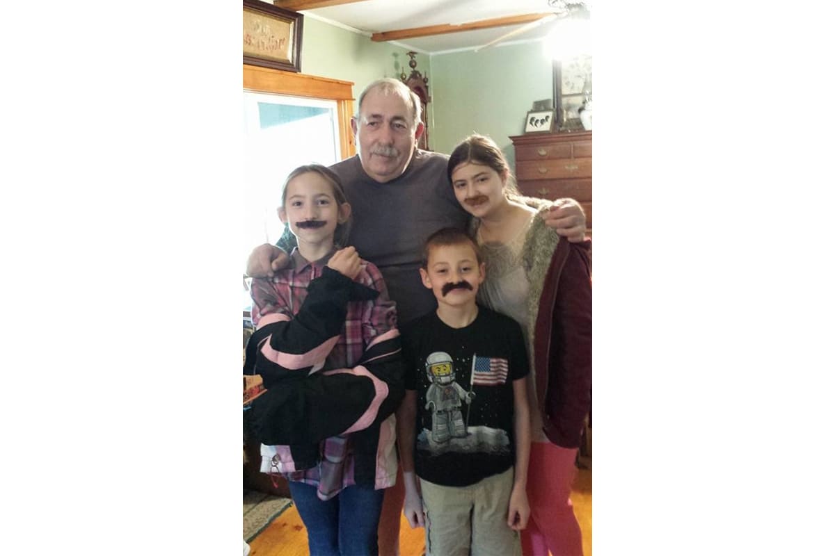 Family of five posing indoors with fake mustaches and smiles, likely playing a playful photo moment.