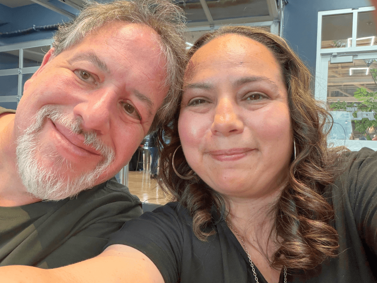 Close-up selfie of a smiling man with a gray beard and a smiling woman with wavy hair indoors.