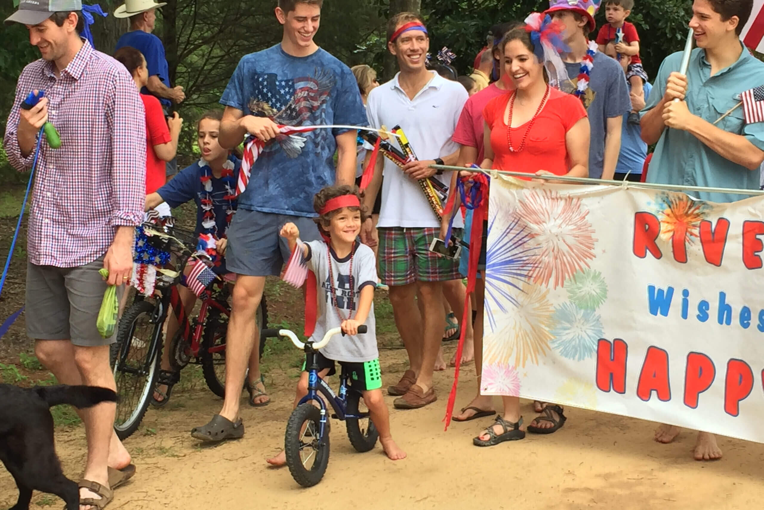 Young boy on a small bicycle at a patriotic parade, surrounded by smiling adults and other kids, with a fireworks banner in the background.