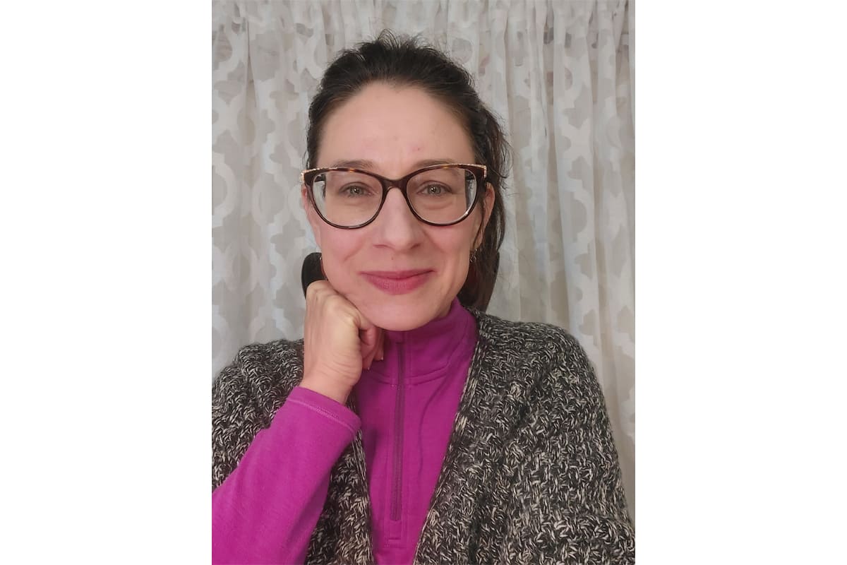 Woman with dark hair and glasses smiling, resting her chin on her hand, wearing a magenta top and grey cardigan area.