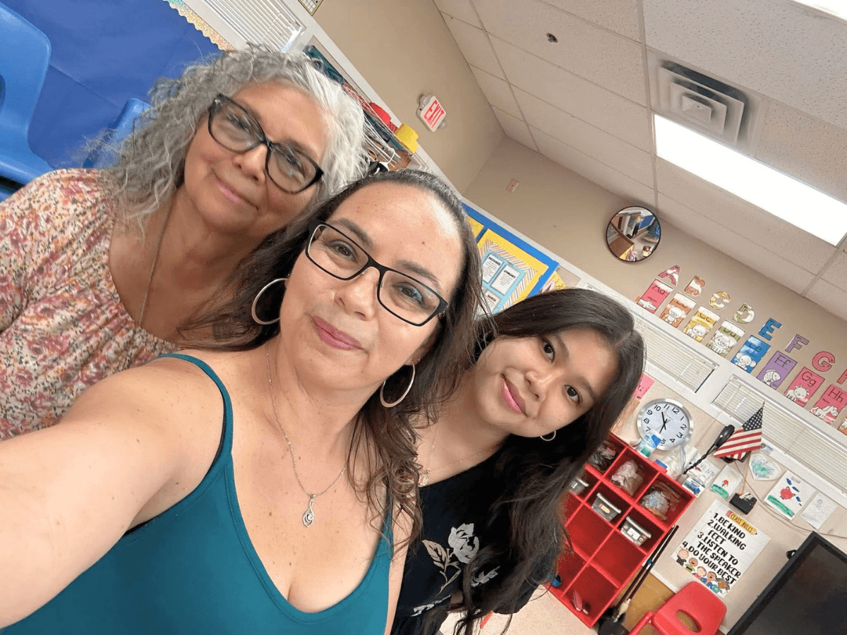 Three women smile for a selfie in a classroom with colorful posters and shelves in the background.