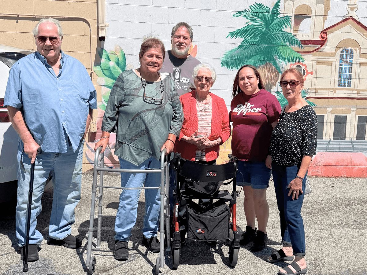 Six adults posing outdoors in front of a colorful mural; two seniors have mobility aids (a walker and a wheelchair) in the foreground.