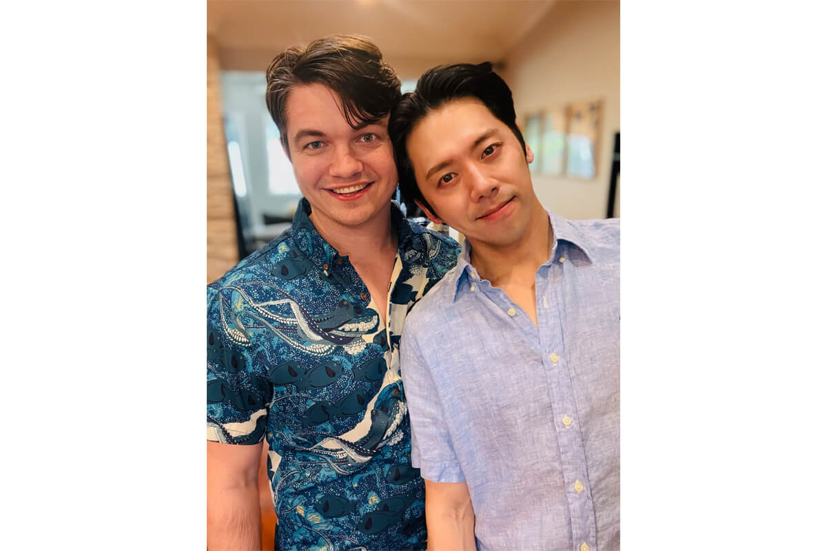 Two men posing together indoors, smiling at the camera; one in a teal patterned shirt and the other in a light blue button-down shirt.