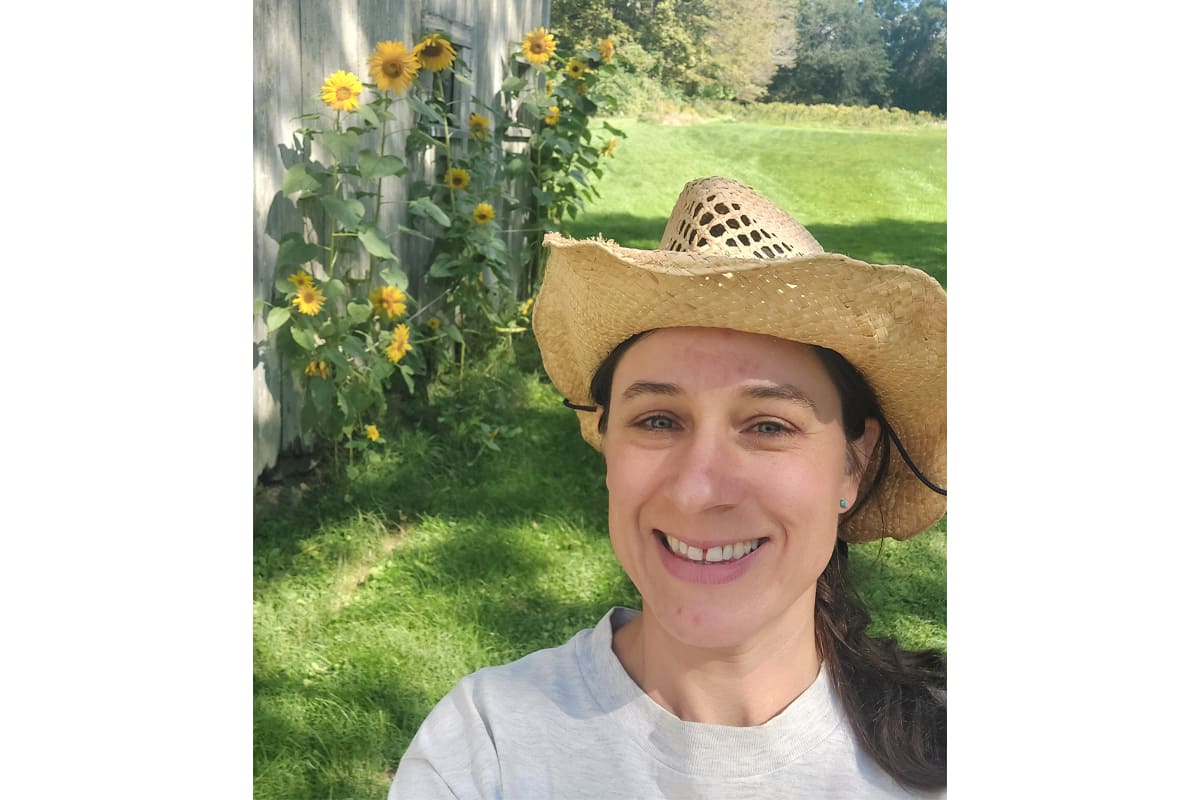 Smiling woman in a straw hat taking a sunny selfie in a yard with sunflowers along a wooden fence