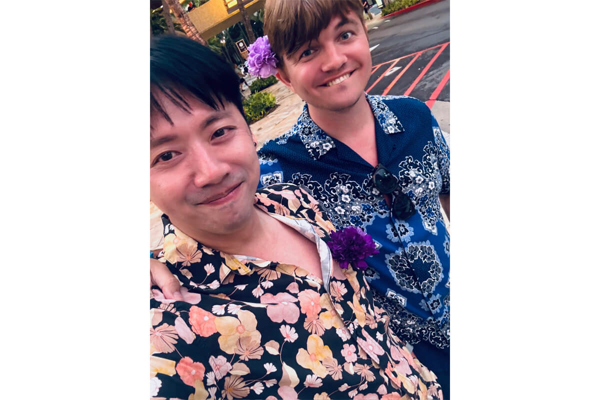 Two men in colorful Hawaiian shirts pose for a selfie outdoors, smiling; one has a purple flower in his hair beneath the camera angle.