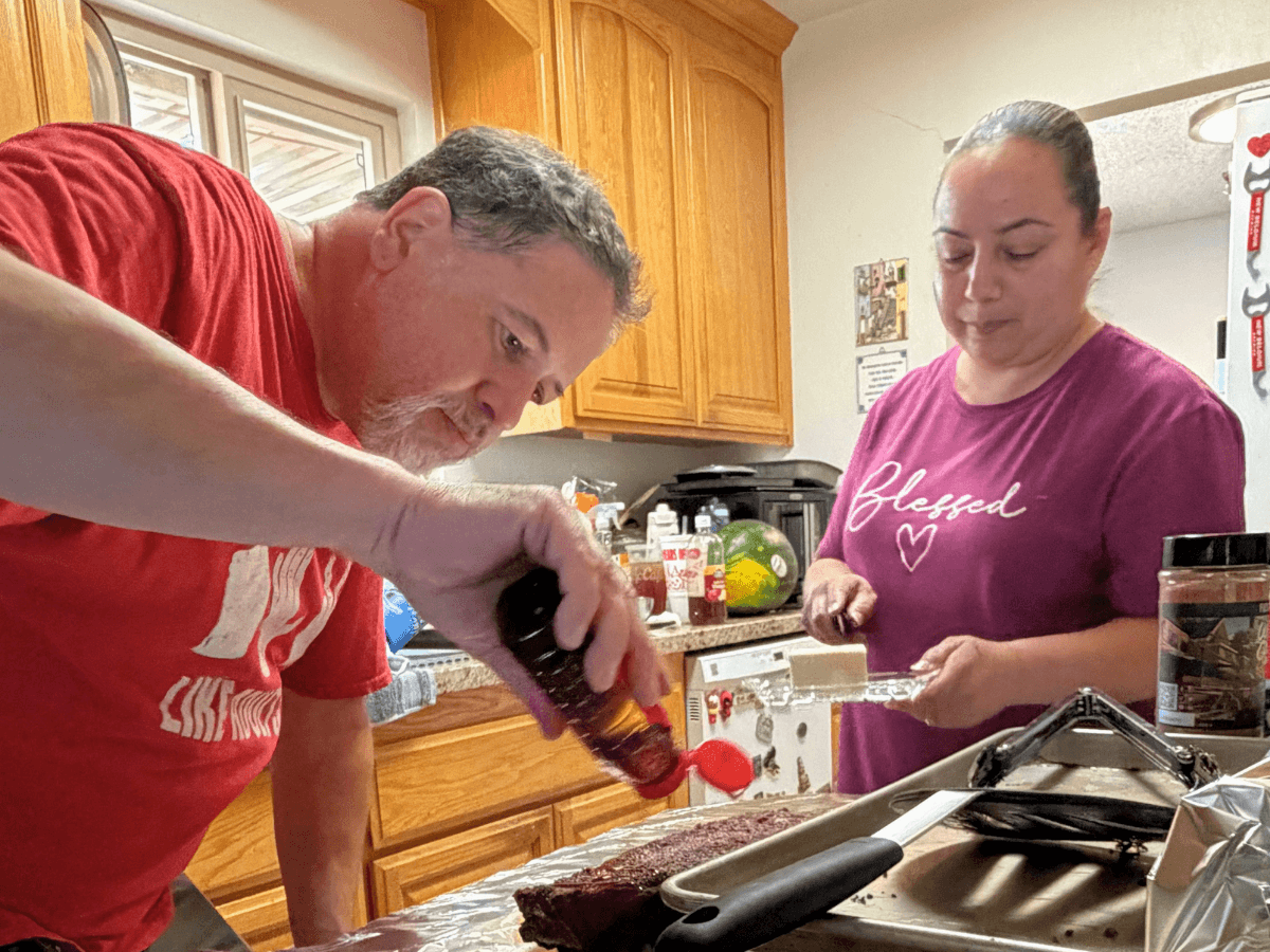 Two people in a kitchen glaze a chocolate brownie on a baking sheet; man pours sauce while woman watches nearby.