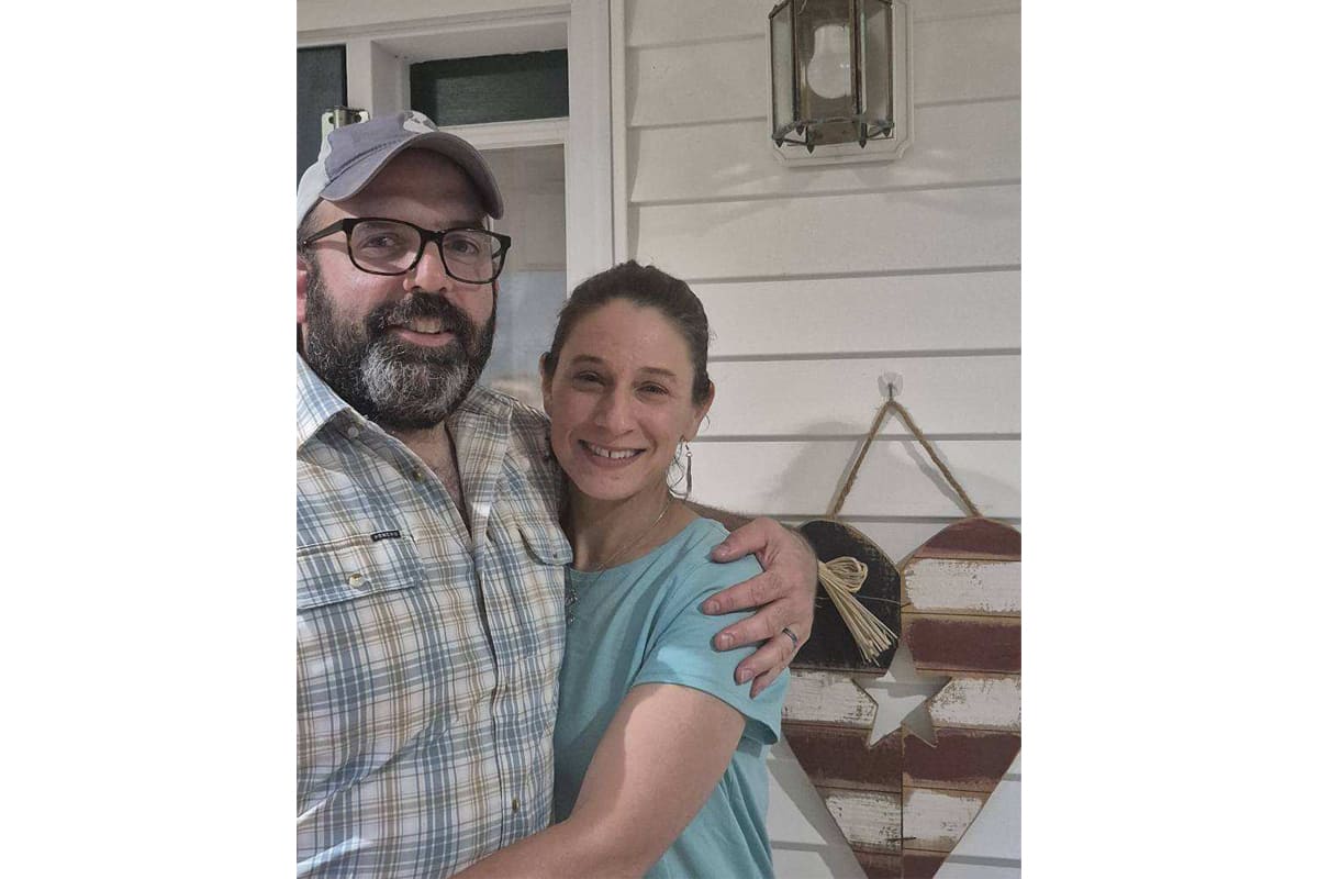 Smiling couple hugs on a porch, man wearing a cap and glasses, woman in a blue shirt beside him.