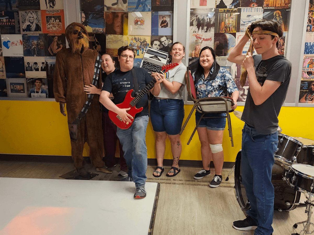 Group of students posing in a music room; a person in a Chewbacca costume stands on the left while a guitarist holds a red electric guitar in front of the group, with others smiling and holding a keyboard, a chair, and a headbanded friend nearby. Drums are visible on the right.
