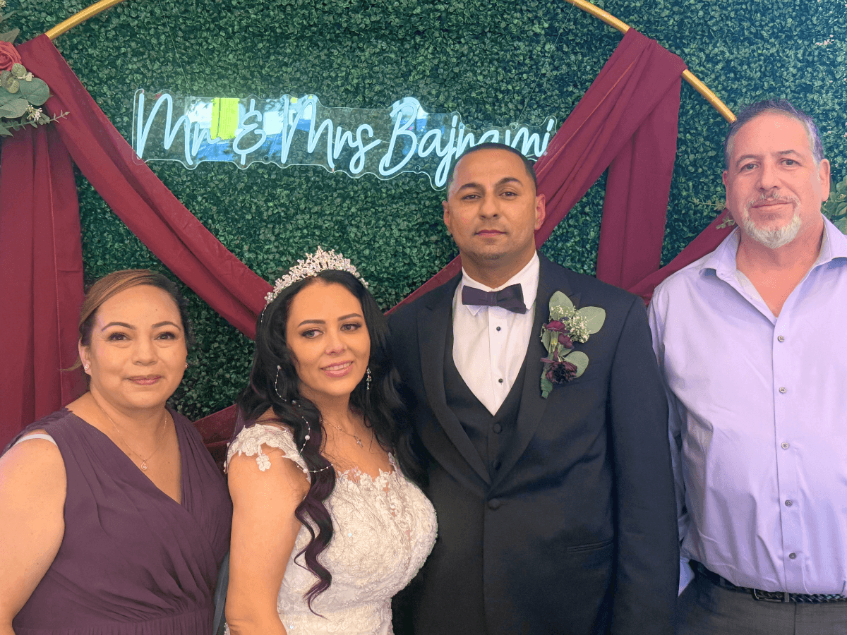 Bride in a lace wedding dress and groom in a black tuxedo pose with two guests against a green hedge backdrop and burgundy drapery at a wedding venue