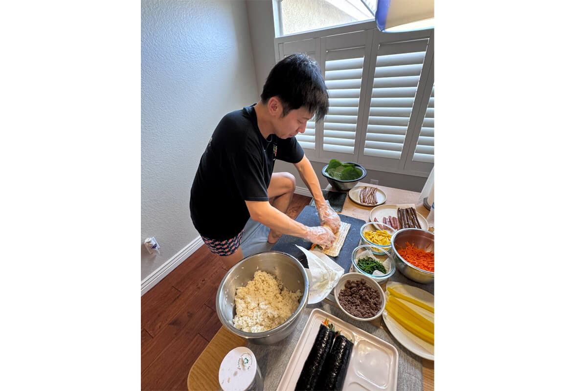 Man in a black shirt rolling a sushi sheet on a table, with bowls of rice, vegetables and seaweed nearby.