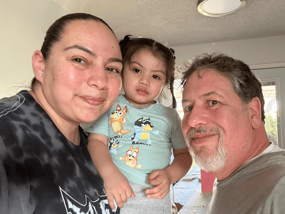 Three people pose for a close selfie indoors: mother on the left, young daughter in the center, and father on the right, all smiling.