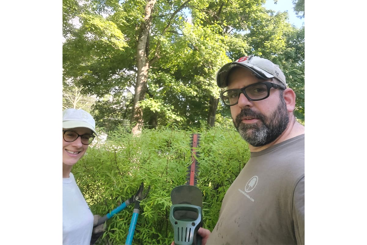 Two adults wearing caps and glasses pose in a garden with bamboo, holding tools (shovel and pruner) for yard work during daytime.