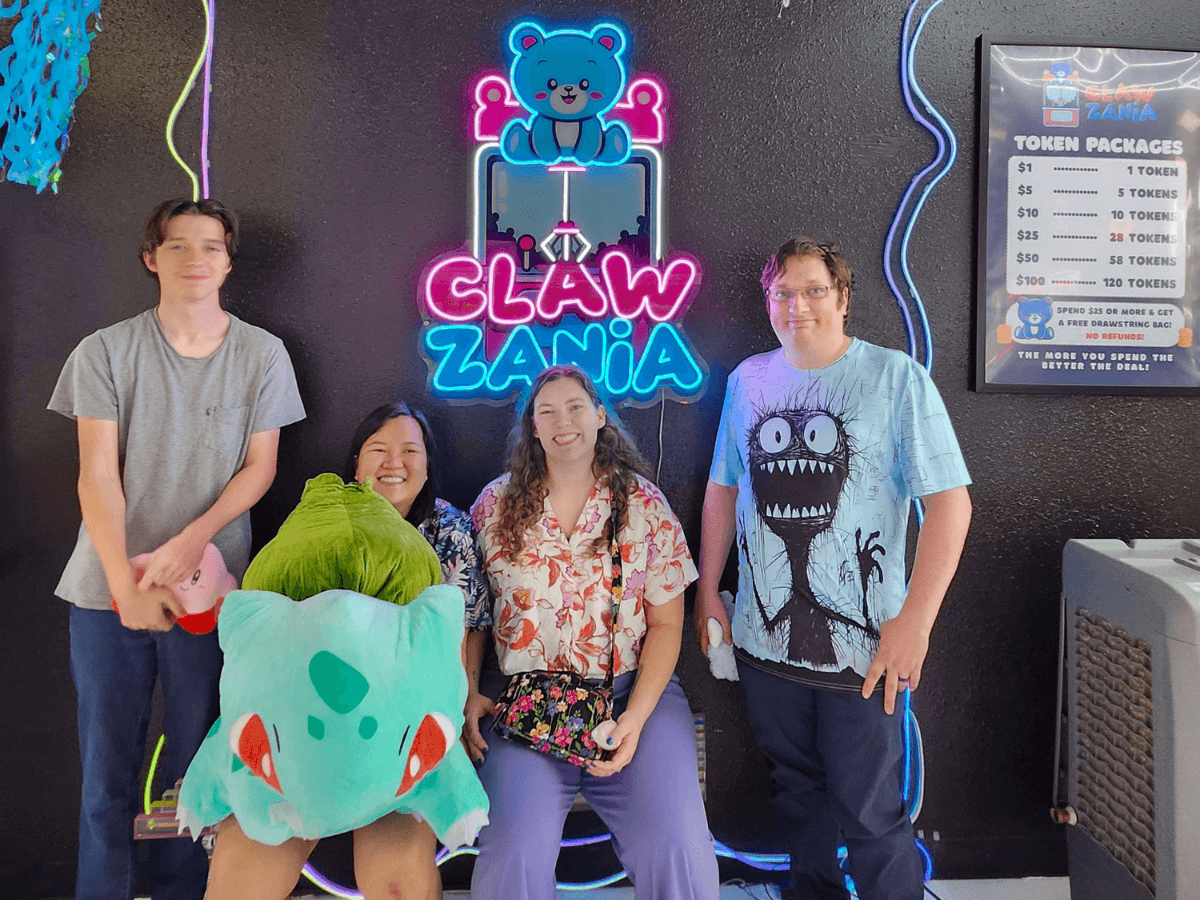 Four people pose in front of a neon 'Claw Zania' sign in an arcade, with a large teal plush toy in the foreground.