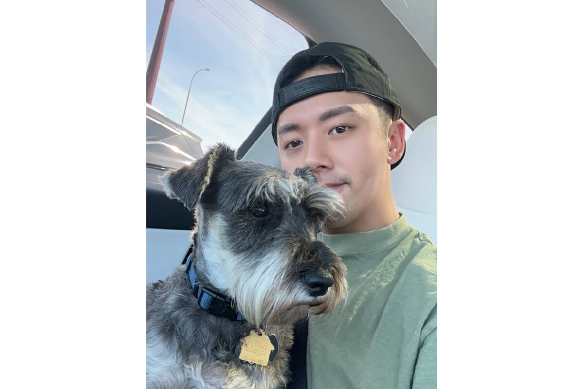 Young man in a backward cap sits in a car with a gray Schnauzer, both facing the camera close up.